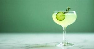 A light green cocktail in a coupe glass, garnished with a cucumber slice and a sprig of rosemary, set against a green background on a marble surface.