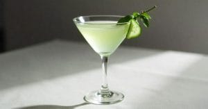 A pale green cocktail in a martini glass, garnished with a cucumber slice and a small leafy sprig, set on a white tablecloth with soft natural light.