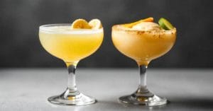 Two yellow-orange cocktails in coupe glasses; the left drink is garnished with banana slices, while the right has a cinnamon rim, citrus slices, and a scoop of ice cream.