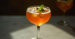A coupe glass filled with an orange cocktail, garnished with a mint sprig and cucumber slice, set on a light surface with another similar drink blurred in the background.